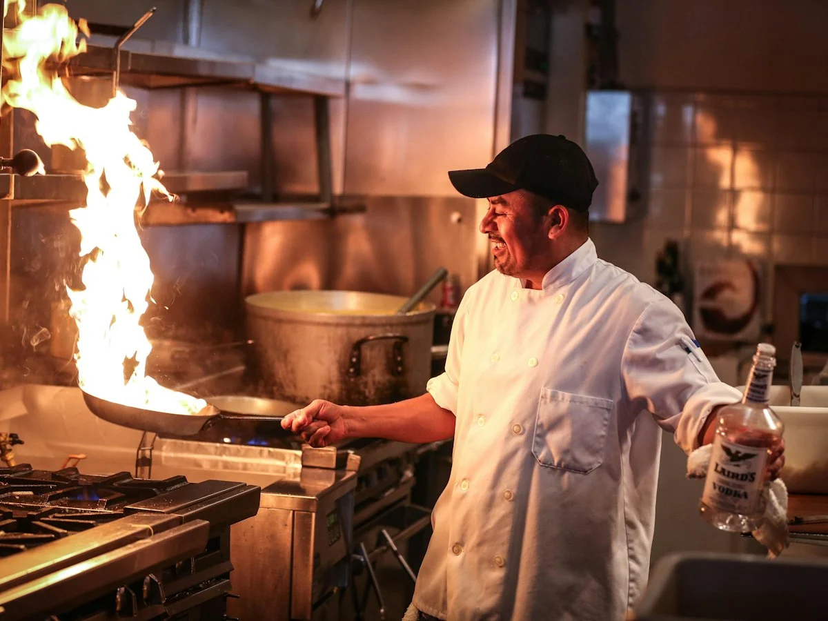 A chef at a commercial range flambéing a dish in a pan